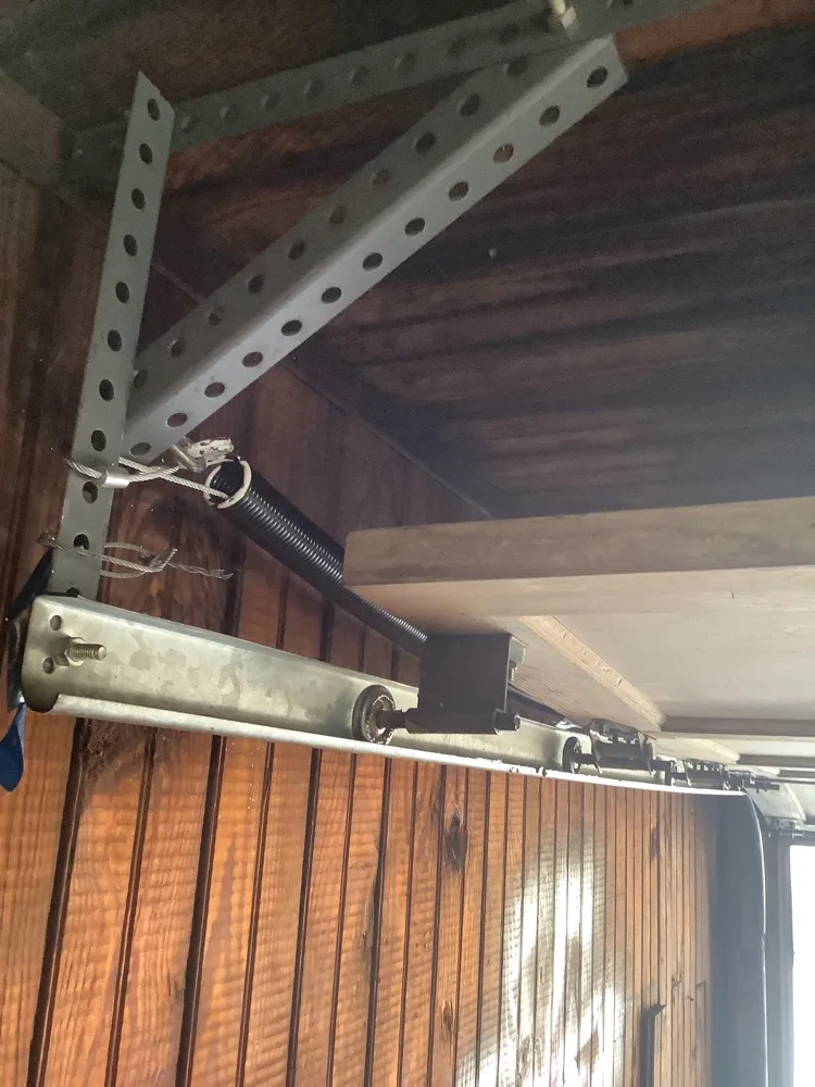 A punched steel hanger bracket carrying the horizontal track above a wood-panelled garage door, with an extension spring and cable ends hooked into it.