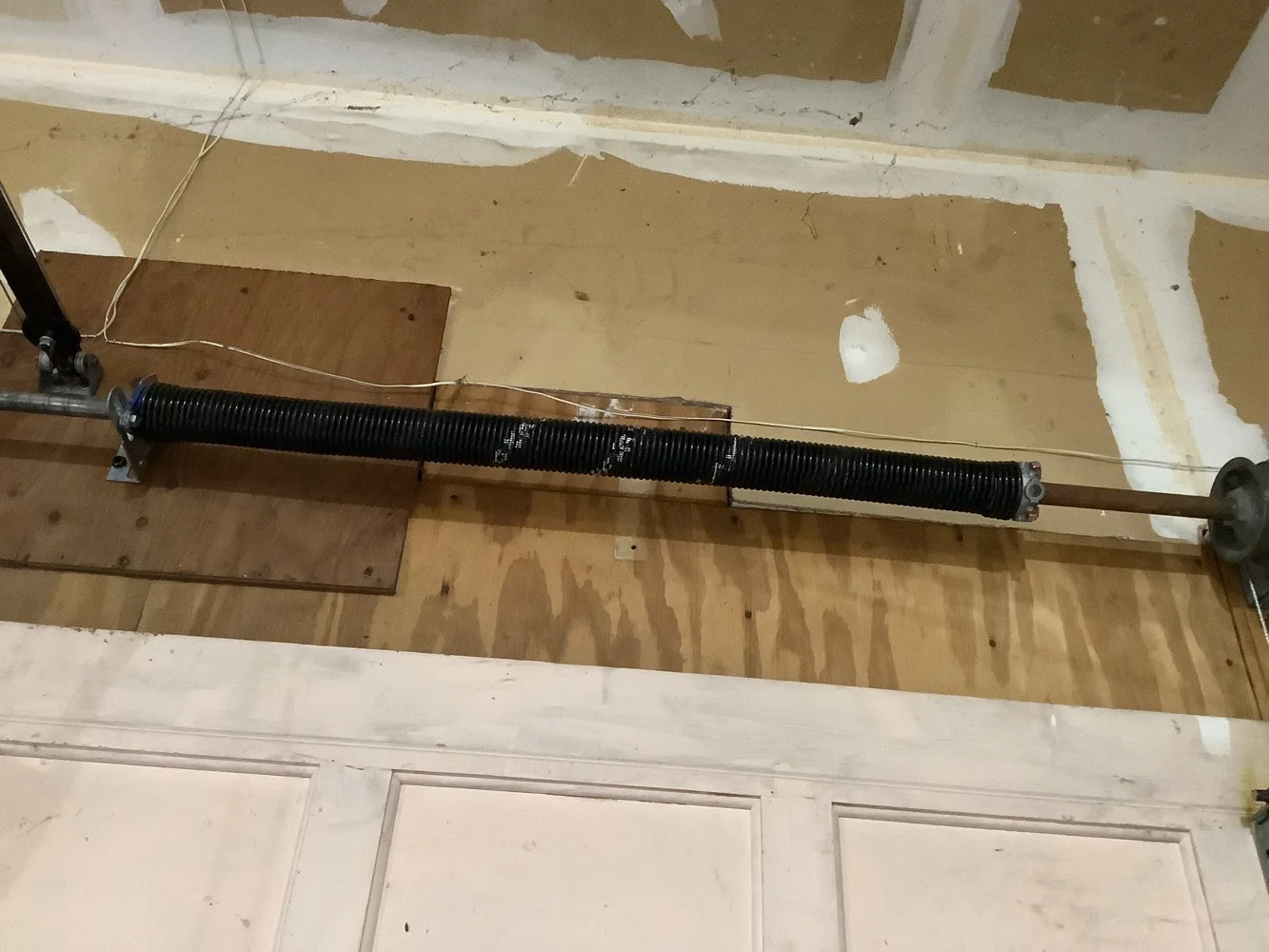 A single black torsion spring wound onto its shaft across a garage header of bare plywood and patched drywall, the cable drum just visible at the far end.