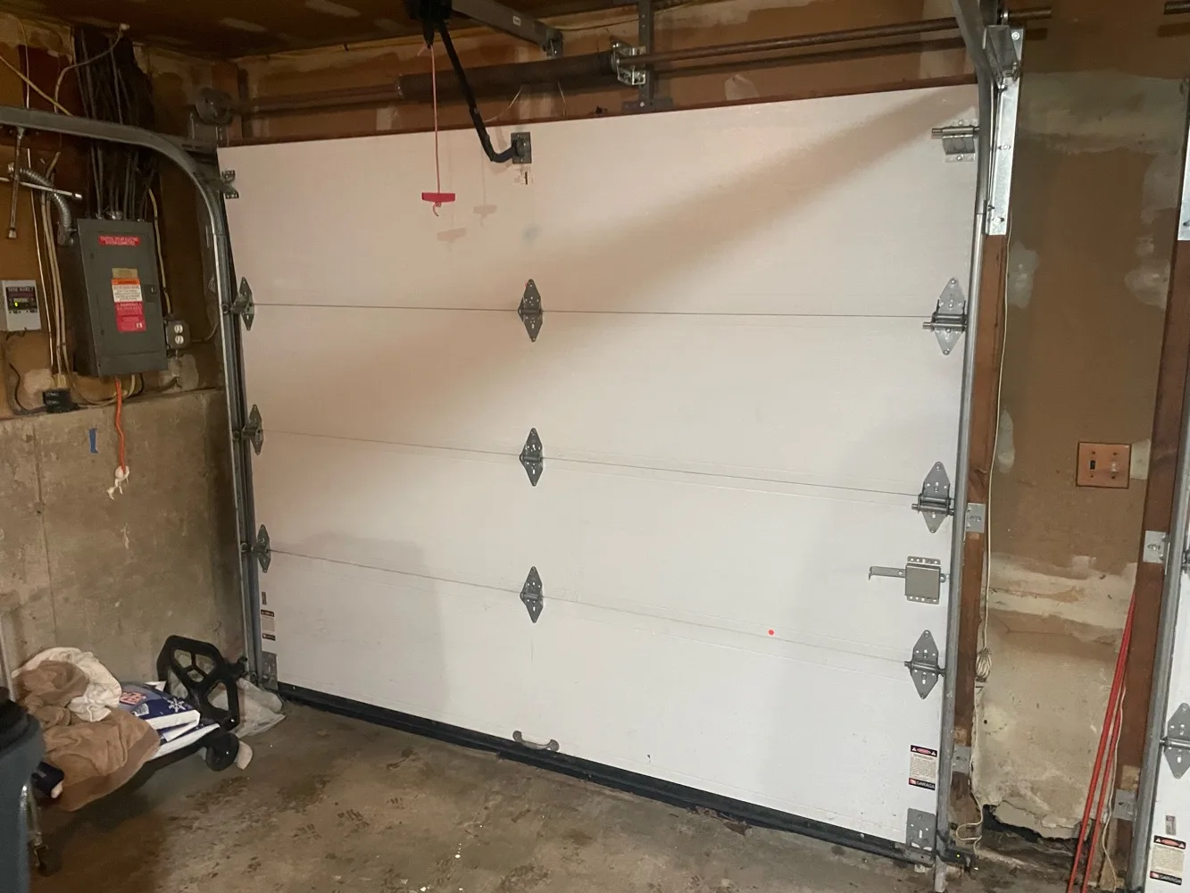 The inside of a single garage with the white sectional door closed, vertical tracks bolted to both jambs, decorative strap hinges on each panel seam and an electrical panel on the wall alongside.