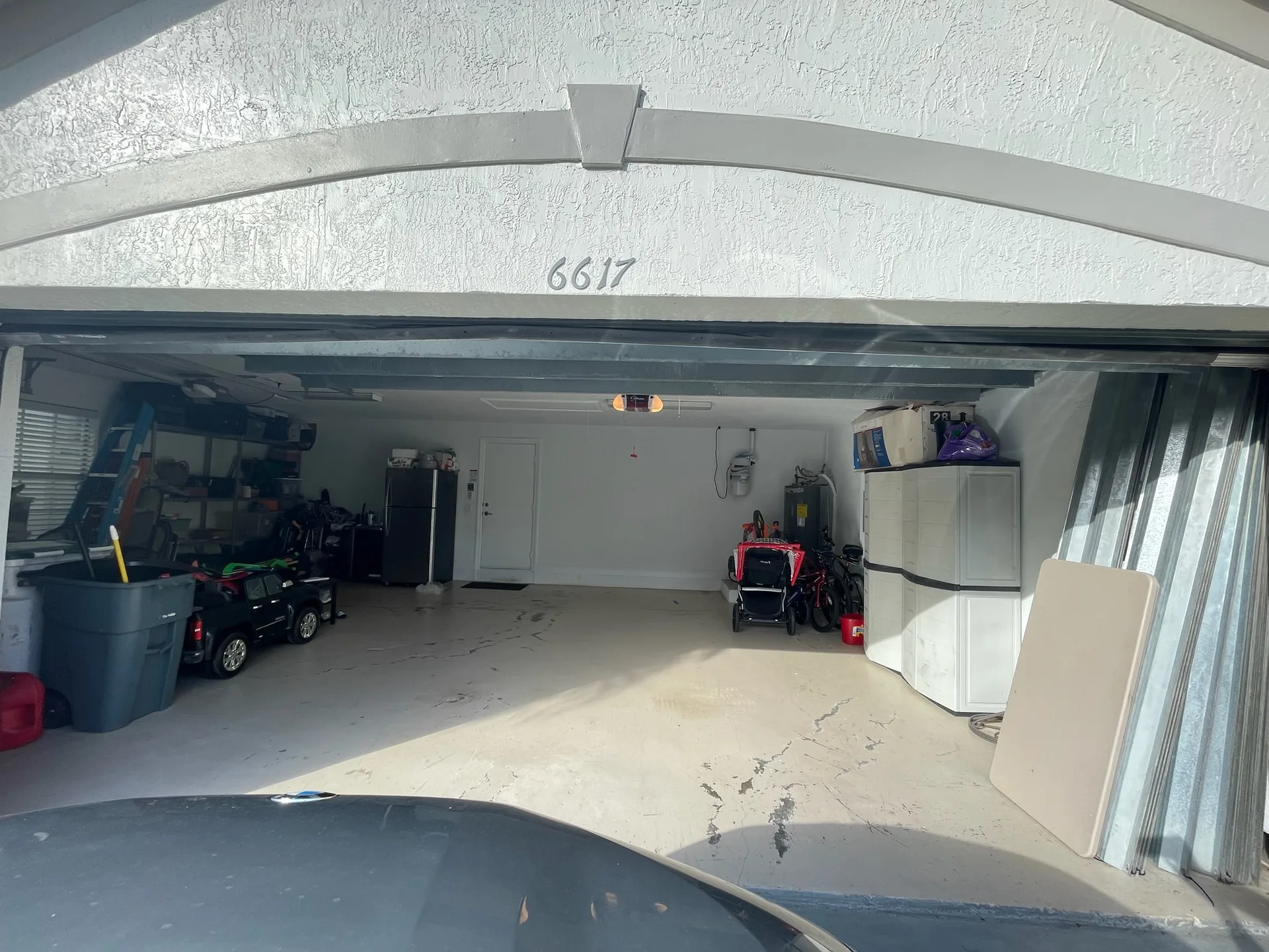 A two-car garage standing fully open beneath a curved stucco arch, hard sunlight falling across the bare slab, with shelving, a ladder and a chest freezer along the side wall.