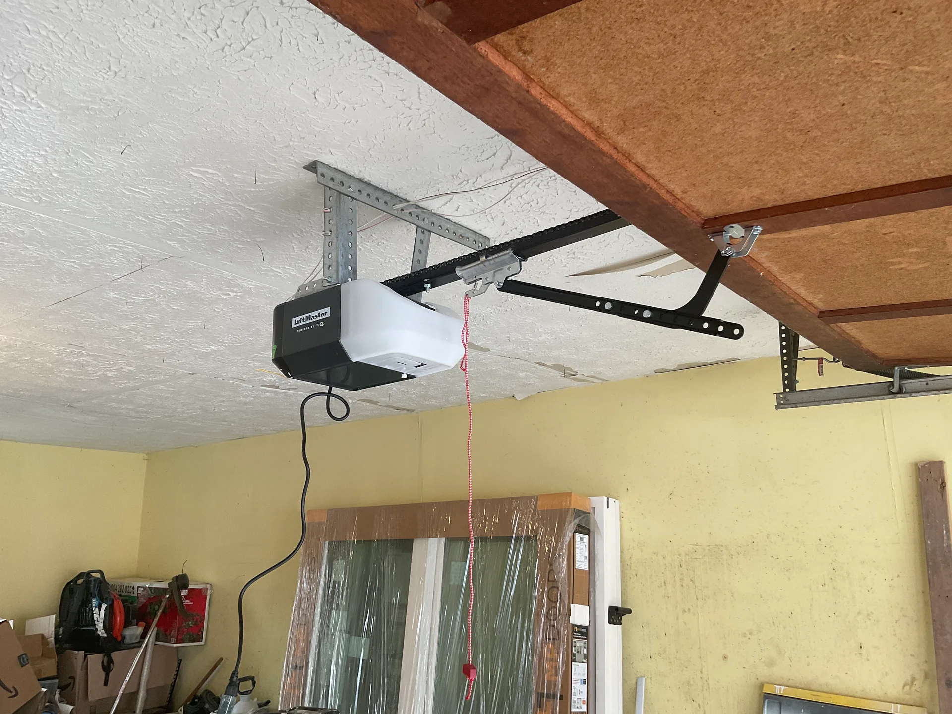A LiftMaster opener head hanging on punched angle iron beneath a textured white ceiling, its rail and door arm running off to the top of the door and the release cord dangling below.